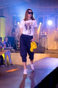 Eine junge Frau präsentiert auf dem Laufsteg ein modernes Streetstyle-Outfit. Sie trägt ein weißes T-Shirt mit einem auffälligen Print – eine stilisierte Katze in Sonnenbrille, Rollkragen und Lederjacke, die einen Bulldoggen-Welpen spazieren führt. Dazu kombiniert sie eine weit geschnittene, knielange, dunkelblaue Hose und weiße Plateau-Sneaker. In der rechten Hand hält sie eine gelbe Trucker-Cap. Ihr Look wird durch eine große, eckige Sonnenbrille, ein schwarzes Perlenhalsband und sichtbare Tattoos am linken Unterarm ergänzt. Der Laufsteg ist von Scheinwerfern beleuchtet, im Hintergrund sind Zuschauer und eine DJ-Anlage zu sehen.