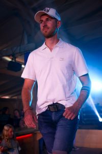 Ein junger Mann mit Vollbart läuft auf einem Laufsteg. Er trägt ein weißes Poloshirt mit kurzem Reißverschluss, eine dunkle Jeansshorts und einen schwarzen Gürtel mit großer Schnalle. Auf dem Kopf trägt er eine weiße Baseballkappe mit einem schwarzen Emblem. Sein rechter Arm ist mit einem schwarzen Armband und einem auffälligen schwarzen Tattoo mit drei parallelen Streifen geschmückt. Im Hintergrund ist Bühnenbeleuchtung mit blauen und lila Scheinwerfern zu sehen, sowie einige Zuschauer, darunter ein blondes Kind links im Bild.