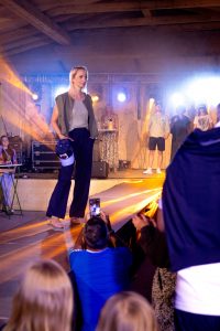 Eine blonde Frau läuft elegant über den Laufsteg. Sie trägt eine ärmellose, olivgrüne Weste über einem hellgrauen, gerippten Top, kombiniert mit einer weit geschnittenen, dunkelblauen Hose. In der rechten Hand hält sie eine sportliche Cap in Blau und Weiß, was dem Outfit eine moderne, lässige Note verleiht. Ihr Look wird durch flache Sandalen abgerundet. Im Hintergrund sind weitere Models und Zuschauer zu sehen, während das Licht in warmen Gelb- und Blautönen für eine stimmungsvolle Atmosphäre sorgt.