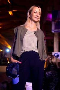 Eine lächelnde Frau posiert auf dem Laufsteg und strahlt Selbstbewusstsein aus. Sie trägt ein ärmelloses, olivgrünes Blouson über einem gerippten Tanktop in passendem Farbton. Die Hose ist elegant, dunkel und weit geschnitten – ein moderner, sportlich-urbaner Look. In der Hand hält sie eine dunkelblaue Cap, was dem Outfit einen lässigen Touch verleiht. Warmes Licht und neugierige Blicke aus dem Publikum unterstreichen die entspannte Atmosphäre des Abends.