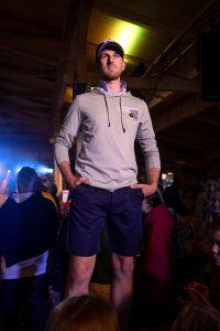 Ein junger Mann steht selbstbewusst auf einem Podest oder Laufsteg in einem Veranstaltungsraum mit Holzbalkendecke. Er trägt eine dunkelblaue Baseballkappe, einen hellgrauen Hoodie mit Aufschrift auf der Brust sowie dunkelblaue Shorts. Die Hände hat er locker in die Hosentaschen gesteckt. Im Hintergrund erkennt man zahlreiche Zuschauer:innen, die das Geschehen interessiert verfolgen. Die Lichtstimmung ist dramatisch mit violett-blauen Spotlights, die von der Bühne ausgehen und das Model von hinten beleuchten.