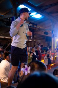 Ein junger Mann mit Basecap und hellem Hoodie steht in einem Veranstaltungssaal auf einer erhöhten Fläche. Er trägt dunkelblaue Shorts und hält mit einer Hand seinen Hoodie-Kragen fest. Der Hoodie hat eine kleine, helle Aufschrift auf der Brust. Um ihn herum sitzen und stehen viele Zuschauer:innen, darunter Kinder und Erwachsene, die aufmerksam die Modenschau verfolgen. Das Licht ist blau-violett und stimmungsvoll, was die Atmosphäre festlich wirken lässt. Im Vordergrund sind unscharf die Köpfe weiterer Zuschauer:innen zu erkennen, darunter ein Mädchen mit einer lilafarbenen Blume im Haar.