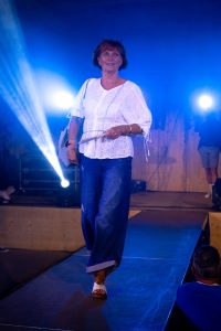 Eine Frau läuft selbstbewusst über den Laufsteg, gekleidet in eine weiße Bluse mit Lochstickerei und weiten, dunkelblauen Jeans mit umgeschlagenem Saum. In der Hand hält sie einen Fächer, über der Schulter trägt sie eine hellgraue Tasche. Ihre weißen Sandalen runden das luftige Sommeroutfit ab. Die Szene ist von intensivem blauen Licht und Bühnenstrahlern umgeben, was dem Auftritt eine dynamische und sommerliche Atmosphäre verleiht.