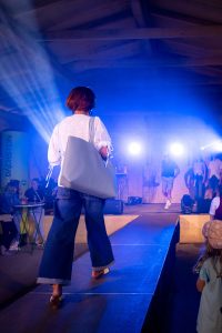 Eine Frau mit kurzen braunen Haaren läuft auf einem Laufsteg von hinten gesehen. Sie trägt eine luftige, weiße Bluse mit Lochstickerei, eine weite Jeans mit Umschlag am Saum und weiße Sandalen mit brauner Sohle. Über der Schulter trägt sie eine große, hellgraue Schultertasche im Shopper-Stil. Die Lichtstimmung ist blau-violett, im Hintergrund sind Zuschauer, weitere Models und starke Scheinwerfer auf der Bühne zu sehen. Ein Kind mit Mütze schaut von der Seite interessiert zu.