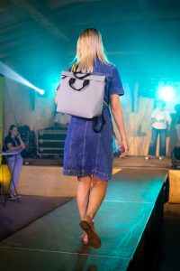 Eine Frau mit blondem, schulterlangem Haar geht über einen Laufsteg. Sie trägt ein kurzes Jeanskleid mit kurzen Ärmeln und eine beigefarbene Sandale. Auf dem Rücken hat sie einen großen, eckigen, hellgrauen Rucksack mit schwarzen Trägern und einem gelben Detail. Die Bühne ist in türkisfarbenes Licht getaucht, und im Hintergrund sind weitere Personen sowie technische Bühnenausstattung zu sehen.