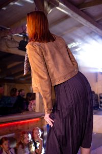Eine Frau steht auf einem Laufsteg mit dem Rücken zur Kamera und blickt leicht zur Seite. Sie trägt eine cognacfarbene Wildlederjacke mit silbernen Nieten an Schultern und Bund sowie einen schwarzen, knielangen Plisseerock. Ein Leopardenmuster-Schal hängt locker aus der Jacke. Im Hintergrund sitzen Kinder und Erwachsene in der ersten Reihe des Publikums, einige Kinder schauen staunend zur Frau hoch. Die Szene wird von violettem und weißem Bühnenlicht erhellt, das durch das Dach einer Holzhütte strahlt.