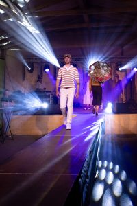 Ein Mann läuft selbstbewusst auf einem beleuchteten Laufsteg bei einer Indoor-Modenschau. Er trägt ein kurzärmeliges Poloshirt mit Retro-Ringelstreifen in Orange, Grün, Beige und Weiß, eine beige Hose und weiße Sneakers. Abgerundet wird sein Look mit einer braunen Trucker-Cap mit Patch. Im Hintergrund laufen weitere Models, darunter eine Person mit einem auffälligen Regenschirm im Leopardenmuster und rosa Schrift. Die Bühne ist mit Spots und Nebellicht atmosphärisch in Szene gesetzt. Die Lichtstrahlen fächern sich dekorativ zur Seite auf.