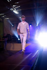 Ein Mann geht über einen Laufsteg bei einer Modenschau in einer Halle mit Holzbalken und Lichtstrahlern. Er trägt ein gestreiftes Poloshirt mit kurzen Ärmeln in den Farben Weiß, Orange, Grün und Beige sowie eine helle, gerade geschnittene Hose. Auf dem Kopf trägt er eine beige Trucker-Cap mit Aufnäher. Der Hintergrund ist mit Bühnentechnik, Musikern und weiteren Models belebt. Die Beleuchtung erzeugt kräftige Lichtstrahlen, die nach vorne strahlen. Das Model geht mit festem Blick und sicherem Gang direkt auf das Publikum zu.