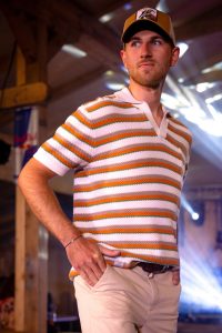 Ein junger Mann läuft über den Laufsteg bei einer Indoor-Modenschau. Er trägt ein kurzärmeliges, gestricktes Poloshirt mit Streifen in Orange, Weiß, Grün und Beige. Dazu kombiniert er eine helle beigefarbene Stoffhose mit braunem Gürtel. Auf dem Kopf trägt er eine beige-braune Trucker-Cap mit einem aufgenähten Outdoor-Patch. Die Kulisse ist eine holzvertäfelte Halle mit Scheinwerferlicht, das von der Decke strahlt. Der Mann blickt selbstbewusst zur Seite und hat beide Hände in den Hosentaschen.