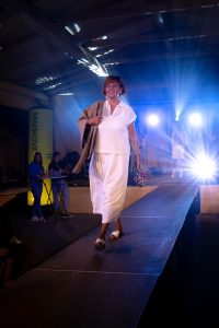 Eine ältere Frau schreitet lächelnd über den Laufsteg bei einer Modenschau in einer Holzhalle mit Bühnenbeleuchtung. Sie trägt ein luftiges, weißes Outfit bestehend aus einer weiten, fließenden Hose und einem ärmellosen Strick-Oberteil mit Kragen. Über der Schulter liegt eine beige Wildlederjacke. Ihre Accessoires sind dezent, in der rechten Hand hält sie einen bunten Regenschirm mit Leopardenmuster. Offene weiße Sandalen und goldene Ohrringe runden das stilvolle Sommer-Ensemble ab. Im Hintergrund sind Zuschauer und andere Models auf der Bühne zu sehen, während Lichtstrahlen die Szene lebendig beleuchten.