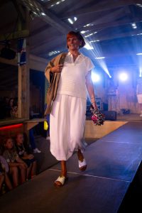 Eine Frau mittleren Alters schreitet elegant über den Laufsteg bei einer Modenschau. Sie trägt ein sommerliches, weißes Ensemble bestehend aus einem gehäkelten Kurzarm-Oberteil und einem luftigen, wadenlangen Rock. Über ihre Schulter liegt lässig eine beigefarbene Jacke. In ihrer rechten Hand hält sie einen zusammengefalteten, leopardenbedruckten Regenschirm mit pinker Schrift. Ihre offenen weißen Sandalen mit goldenen Details runden das Outfit stilvoll ab. Sie lächelt und blickt selbstbewusst in Richtung Publikum, das auf Bänken neben dem Laufsteg sitzt. Lichtstrahlen und Bühnenbeleuchtung erzeugen eine festliche Atmosphäre.