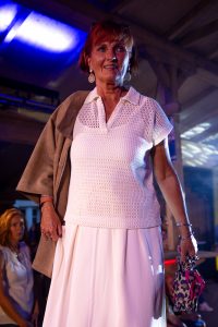 Eine Frau mittleren Alters steht auf einem Laufsteg bei einer Modenschau. Sie trägt ein elegantes, cremeweißes Outfit, bestehend aus einem gehäkelten Kurzarm-Poloshirt und einem weit fallenden, fließenden Rock. Über ihre Schulter hängt eine beigefarbene Jacke, in ihrer Hand hält sie einen zusammengefalteten Leoparden-Regenschirm mit pinker Schrift. Sie trägt auffällige Ohrringe und wirkt selbstbewusst und freundlich. Im Hintergrund erkennt man ein interessiertes Publikum. Die Szene ist atmosphärisch mit farbigem Licht beleuchtet.