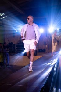 Ein älterer Mann schreitet selbstbewusst über den Laufsteg bei einer Modenschau. Er trägt ein helles Sommeroutfit bestehend aus einem blasslila Poloshirt, einer offenen hellen Jacke, weißen Shorts, weißen Tennissocken und weißen Sneakers. Er hat die Hände lässig in den Hosentaschen und trägt eine dunkle Sonnenbrille. Der Laufsteg ist von blauem Licht und Spots beleuchtet. Im Hintergrund stehen weitere Models, darunter eine Frau mit einem Leoparden-Regenschirm. Die Atmosphäre ist lebendig und sommerlich-modern.