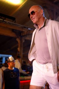 Ein älterer Mann mit Glatze und dunkler Sonnenbrille läuft über einen Laufsteg bei einer Modenschau. Er trägt ein helles Poloshirt, eine offene, weiße Sommerjacke und weiße Shorts. Mit einem Lächeln und der rechten Hand in der Hosentasche wirkt er entspannt und selbstbewusst. Im Hintergrund sind Menschen an einer Bar zu sehen, eine Person trägt ein T-Shirt mit Fahrradaufdruck und eine weiße Kappe. Die Szene ist in warmes Licht getaucht, das von der Holzdecke reflektiert wird.