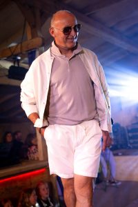 Ein älterer Mann mit Glatze läuft selbstbewusst über den Laufsteg einer Modenschau. Er trägt ein helles Poloshirt, eine weiße, leichte Jacke und weiße Shorts. In der rechten Hand steckt sie lässig in der Hosentasche, die linke hält den Jackensaum. Eine dunkle Sonnenbrille rundet den entspannten Sommer-Look ab. Im Hintergrund sind Zuschauer zu sehen, darunter auch Kinder, die dem Auftritt interessiert folgen. Die Szene ist in warmes Scheinwerferlicht getaucht und spielt sich in einer überdachten Holzkonstruktion ab.