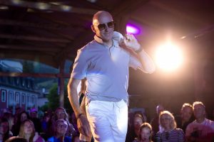 Ein älterer Mann mit Glatze läuft charmant über einen Laufsteg bei einer Modenschau. Er trägt ein helles Poloshirt und eine weiße Hose. Lässig hat er eine weiße Jacke über die rechte Schulter gelegt. Seine dunkle Sonnenbrille und das selbstbewusste Lächeln unterstreichen seine souveräne Ausstrahlung. Im Hintergrund ist das Publikum sichtbar, das aufmerksam und interessiert dem Laufsteggeschehen folgt. Die Szene wird von bunten Scheinwerfern und warmem Bühnenlicht erhellt.