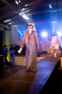 Eine Frau läuft selbstbewusst über einen Laufsteg bei einer Indoor-Modenschau. Sie trägt ein langes Kleid mit Leopardenmuster, eine braune Wildlederjacke mit Nietenverzierungen und dunkle Sonnenbrille. In der rechten Hand hält sie einen gelb-weiß gemusterten, zusammengefalteten Regenschirm. Hinter ihr ist die Bühne mit Lichttechnik und DJ-Pult sichtbar.