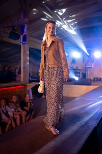Eine Frau geht lächelnd über einen Laufsteg in einer Veranstaltungshalle. Sie trägt ein bodenlanges Sommerkleid mit Leopardenmuster, eine braune Wildlederjacke mit Nietenverzierung und offene Sandalen. In der rechten Hand hält sie einen zusammengefalteten Schirm. Im Hintergrund ist eine Bühne mit DJ-Pult und Scheinwerferlicht zu sehen. Kinder sitzen fasziniert am Rand des Laufstegs und beobachten das Geschehen. Die Atmosphäre ist lebendig und stimmungsvoll.