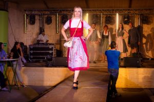 Eine junge Frau mit blonden Haaren steht in Pose auf einem Laufsteg und trägt ein klassisches Dirndl in kräftigem Rot. Die weiße Spitzenbluse ist hochgeschlossen mit kurzen Ärmeln. Die graue Schürze ist mit roten Ornamenten verziert und auf der rechten Seite gebunden. Die Frau trägt schwarze Schuhe mit Absatzriemen und eine kleine geflochtene Umhängetasche mit hellbraunem Deckel. Ihre Hände ruhen selbstbewusst in der Hüfte. Im Hintergrund sind weitere Models in Trachtenmode zu sehen.
