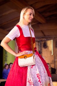 Eine junge Frau mit langen, blonden Haaren trägt ein festliches Dirndl in leuchtendem Rot mit silberfarbenen Knöpfen und einer Schürze in Rot und Weiß mit floralen Mustern. Unter dem Dirndl trägt sie eine weiße Spitzenbluse mit kurzen Ärmeln und Stehkragen. Umgehängt hat sie eine kleine, helle Korbtasche mit braunem Lederdeckel und goldener Schnalle. Die Frau steht selbstbewusst mit einer Hand in der Taille auf einem Laufsteg. Im Hintergrund sind verschwommen weitere Gäste sichtbar.