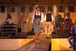 Eine Frau mit kurzen roten Haaren läuft mittig auf einem Laufsteg. Sie trägt ein traditionelles Dirndl mit weißer Bluse, schwarzem Mieder, olivgrüner Schürze und einem floral gemusterten Rock, der unter der Schürze hervorlugt. Dazu kombiniert sie beigefarbene Pumps und eine schwarze Handtasche. Im Hintergrund stehen weitere Models in Trachtenmode auf der Bühne, darunter Männer in Lederhosen und Frauen in Dirndln. Links im Bild steht ein DJ an einem Mischpult, rechts eine Frau im langen Sommerkleid an einem Stehtisch mit Getränken. Die Bühne ist hell ausgeleuchtet mit professioneller Lichttechnik.