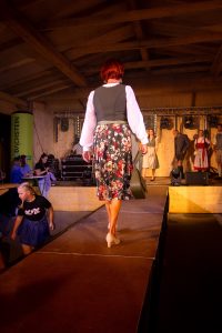 Eine Frau mit kurzen, roten Haaren läuft in einem traditionellen, aber modern interpretierten Trachtenoutfit über den Laufsteg. Sie trägt eine weiße Bluse mit langen Ärmeln, ein dunkelgrünes Mieder und einen knielangen Rock mit farbenfrohem Blumenmuster. Beige Pumps und eine dunkelgrüne Handtasche runden das Outfit ab. Im Hintergrund stehen weitere Models in Trachtenmode auf der Bühne, beleuchtet von Scheinwerfern. Die Atmosphäre wirkt lebendig und festlich.
