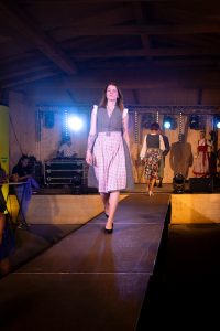 Eine junge Frau schreitet geradeaus auf einem Laufsteg und trägt ein traditionelles Dirndl in dunklem Grün mit feinem Muster, kombiniert mit einer rosa-weiß gemusterten Schürze und einer weißen Bluse mit Rüschenärmeln. Sie hat rotbraunes Haar, trägt schwarze Pumps und hat auffällige Tattoos auf dem linken Arm. Im Hintergrund sieht man weitere Models in Trachtenkleidung auf der Bühne sowie ein DJ-Pult. Die Szenerie ist von Scheinwerfern hell ausgeleuchtet und vermittelt eine festliche Stimmung mit Fokus auf traditioneller Mode.