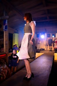 Eine junge Frau läuft über einen Laufsteg und trägt ein traditionelles Dirndl in dunklem Grün mit dezentem Muster, kombiniert mit einer weißen Bluse mit Puffärmeln und einer rosafarben gemusterten Schürze. Sie trägt schwarze Pumps mit Blockabsatz. Ihr rotbraunes Haar fällt glatt auf die Schultern. Im Hintergrund steht ein Mann in Tracht auf der Bühne, links im Bild beobachten Kinder und Erwachsene das Geschehen. Die Laufstegszene ist von buntem Licht ausgeleuchtet, die Atmosphäre wirkt festlich und publikumsnah.