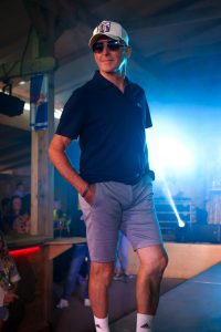 Ein älterer Mann schreitet selbstbewusst über einen Laufsteg. Er trägt ein dunkelblaues Poloshirt, hellgraue Shorts, weiße Adidas-Socken und weiße Sneaker. Ergänzt wird der Look durch eine weiße Baseballcap mit schwarzem Logo und eine dunkle Sonnenbrille. Seine Hände hat er lässig in die Hosentaschen gesteckt. Die Lichtstimmung ist intensiv mit blauem Bühnenlicht im Hintergrund. Im Publikum sitzen mehrere Zuschauer, ein Red-Bull-Aufsteller hängt an einem Holzbalken. Die Szenerie vermittelt eine entspannte, aber stilvolle Sommeratmosphäre.