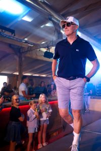 Ein älterer Mann geht selbstbewusst auf einem Laufsteg. Er trägt ein dunkelblaues Poloshirt, hellgraue Shorts, weiße Sneakers mit Sportsocken und eine weiße Baseballkappe mit Logo. Eine schwarze Sonnenbrille unterstreicht seinen lässigen Stil. Seine Hände hat er entspannt in die Hosentaschen gesteckt. Im Hintergrund beobachten Kinder und Erwachsene gespannt das Geschehen, darunter drei Mädchen in Sommerkleidung, die Getränke in der Hand halten. Die Szene ist von blauem und violettem Bühnenlicht durchzogen und vermittelt eine lockere, modische Atmosphäre.