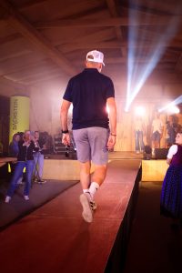 Ein Mann läuft mit dem Rücken zur Kamera auf einem Laufsteg in einer Veranstaltungshalle. Er trägt ein dunkelblaues Poloshirt, hellgraue Shorts, weiße Sportsocken und weiße Sneaker mit schwarzem Muster. In der rechten Hand hält er eine Sonnenbrille, auf dem Kopf trägt er eine weiße Mesh-Cap. Die Laufstegszene ist von starkem Bühnenlicht und Spotstrahlern beleuchtet. Im Hintergrund stehen mehrere Models auf der Bühne, darunter auch Frauen in Sommeroutfits. Zuschauer am Rand fotografieren den Auftritt. Rechts im Bild trägt eine Frau ein traditionelles Dirndl. Die Atmosphäre wirkt modern, locker und lebendig.