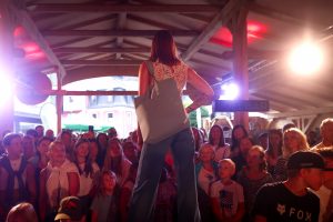 Eine Frau steht auf einem Laufsteg mit dem Rücken zur Kamera und schaut in die Menge. Sie trägt ein ärmelloses, cremefarbenes Spitzentop mit floralem Muster, eine weite Jeans und eine große, hellblaue Tragetasche über der Schulter. Ihr linker Arm ist tätowiert. Sie steht mit einer selbstbewussten Haltung – eine Hand in der Hüfte. Vor ihr steht ein großes, dicht gedrängtes Publikum aus Frauen, Männern und Kindern, das gespannt und lächelnd zur Bühne blickt.
