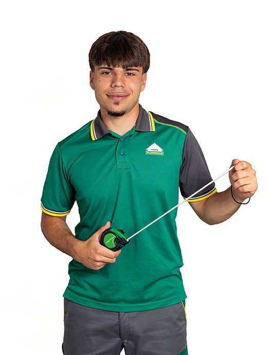 Baustofffachberater Ein junger Mann trägt ein grünes Poloshirt mit grauen und gelben Akzenten sowie eine graue Arbeitshose. Er hält ein grünes Maßband in den Händen, dessen Band er herausgezogen hat, und blickt lächelnd in die Kamera.
