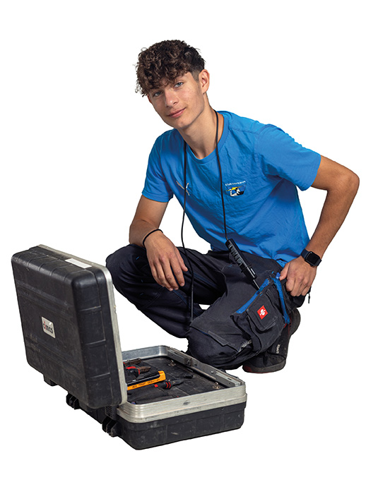 Lehrling_Elektro Ein junger Mann mit lockigem Haar trägt ein blaues T-Shirt und dunkle Arbeitshose. Er kniet neben einem geöffneten schwarzen Werkzeugkoffer, in dem Werkzeuge liegen, und schaut freundlich in die Kamera.