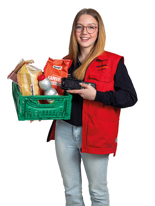 Eine junge Frau mit langen blonden Haaren, Brille und Jeans trägt eine rote Eurogast-Weste. Sie hält eine grüne Transportkiste mit Lebensmitteln, darunter Kaffee, Mehl und Milch, sowie ein elektronisches Handgerät in der anderen Hand. Sie lächelt in die Kamera.