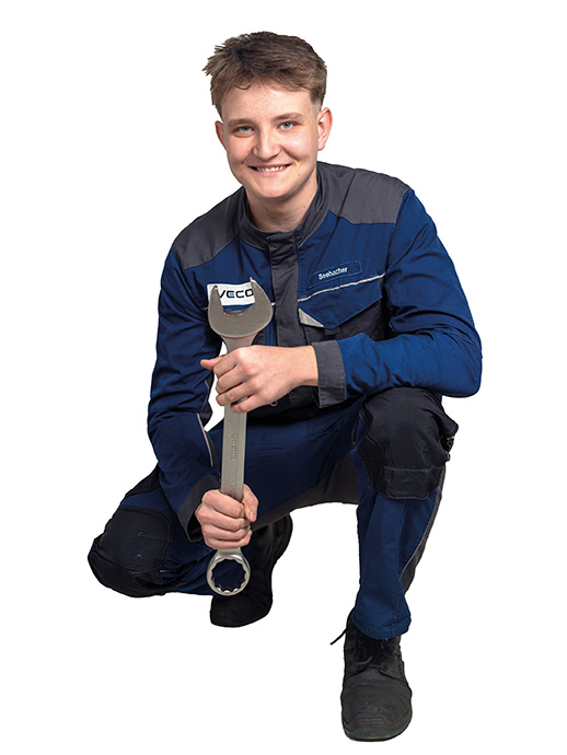 Lehrling KFZ Ein junger Mann in blauer Arbeitskleidung kniet auf einem Bein und hält einen großen Schraubenschlüssel in den Händen. Er lächelt freundlich in die Kamera.