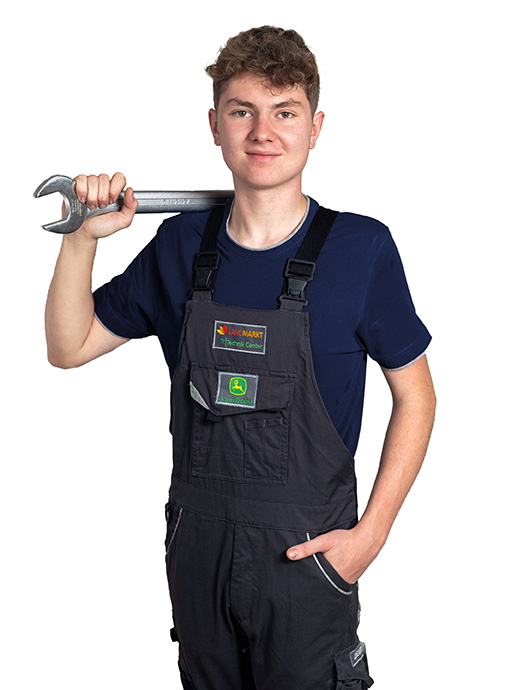Lehrling Landmaschinentechniker Ein junger Mann trägt ein dunkelblaues T-Shirt und eine schwarze Latzhose mit den Logos von Landmarkt Technik Center und John Deere. Er hält einen großen Schraubenschlüssel lässig über die Schulter und schaut selbstbewusst in die Kamera.
