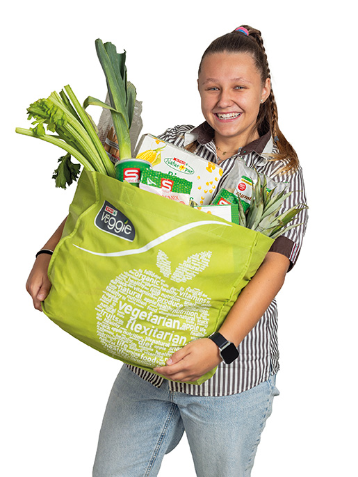 Lehrling_Supermarkt Eine junge Frau mit geflochtenem Pferdeschwanz trägt eine gestreifte Arbeitsbluse und Jeans. Sie hält eine große grüne Einkaufstasche mit dem Aufdruck „veggie – vegetarian – flexitarian“, die mit frischem Gemüse, Obst und verschiedenen Lebensmitteln gefüllt ist. Sie lächelt fröhlich in die Kamera.