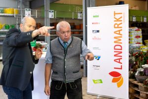 Zwei Männer in traditioneller Tracht stehen im Landmarkt Lagerhaus Gröbming und zeigen in dieselbe Richtung. Im Hintergrund sind Regale mit Gartenzubehör und ein Aufsteller mit dem Landmarkt-Logo sowie den Partnerfirmen SPAR, L&M Fashion, Steinhuber, Elektromann und Eurogast zu sehen.