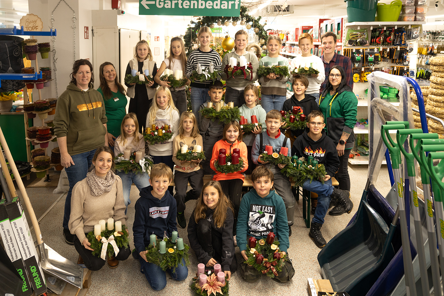 Gruppenfoto im Landmarkt Lagerhaus Aigen: Volksschulkinder der 4a aus Aigen stehen und sitzen gemeinsam mit Mitarbeiter*innen im Markt und halten ihre selbstgebundenen Adventkränze mit Kerzen in die Kamera; im Hintergrund sieht man Regale mit Garten- und Haushaltsbedarf sowie ein Schild „Gartenbedarf“.