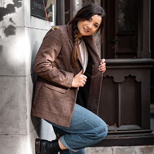 Blazer Outfit Eine junge Frau lehnt lächelnd an einer hellen Steinwand im Außenbereich. Sie trägt einen braunen Blazer, ein helles Oberteil, blaue Jeans und schwarze Stiefeletten. Ihre langen dunklen Haare sind seitlich zu einem Zopf geflochten. Mit einer Hand hält sie den Blazer leicht geöffnet und blickt freundlich in die Kamera. Im Hintergrund sind eine dunkle Holztür und herbstliche Schatten zu sehen.