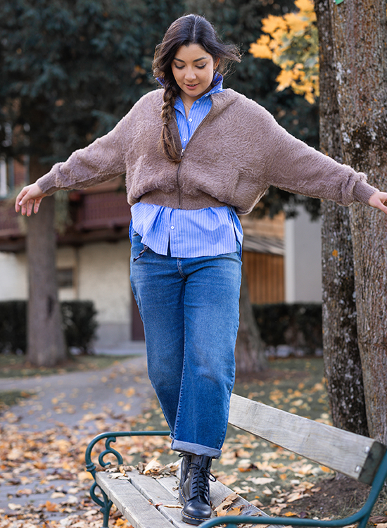 Fake Felljacke Eine junge Frau balanciert mit ausgestreckten Armen auf der Lehne einer Parkbank in einem herbstlichen Park. Sie trägt eine blaue gestreifte Bluse, darüber eine braune, flauschige Strickjacke und eine gerade geschnittene Jeans mit umgeschlagenen Hosenbeinen. Dazu trägt sie schwarze Schnürstiefel. Um sie herum liegen herabgefallene Blätter, und im Hintergrund sind Bäume und ein Haus zu sehen.
