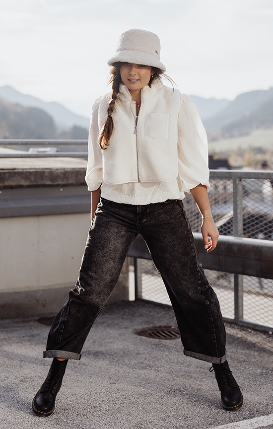 OShape - neue Formen Eine junge Frau steht im Freien auf einer Dachterrasse mit Bergen im Hintergrund. Sie trägt eine weiße Bluse, eine kurz geschnittene weiße Fleeceweste und eine schwarze O-Shape Jeans mit umgeschlagenen Hosenbeinen. Dazu trägt sie schwarze Schnürstiefel und einen hellen, flauschigen Fischerhut. Ihr Haar ist seitlich zu einem Zopf geflochten, und sie blickt lächelnd in die Kamera.