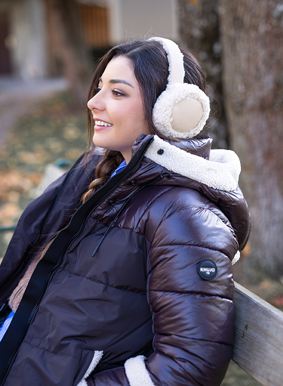 Kopfhörer mit Teddy Eine junge Frau sitzt auf einer Parkbank und lächelt. Sie trägt eine glänzende dunkelbraune Steppjacke mit flauschigem Innenfutter und beige Ohrenschützer aus Fellimitat. Ihr Haar ist seitlich zu einem Zopf geflochten. Im Hintergrund sind herbstliche Bäume und Laub auf dem Boden zu sehen.