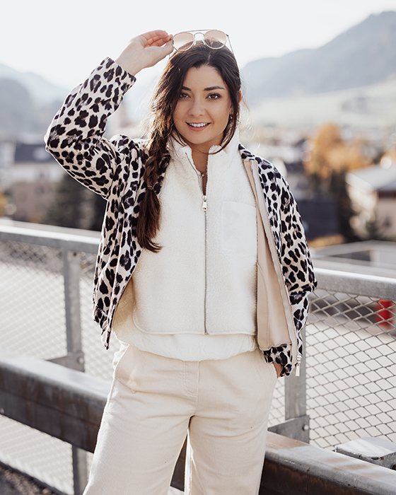 Leo Eine junge Frau steht im Freien auf einer Brücke mit Bergkulisse im Hintergrund. Sie trägt eine helle Hose, eine weiße Fleecejacke und eine Jacke mit Leopardenmuster. Ihre langen, dunklen Haare sind seitlich zu einem Zopf geflochten. Sie lächelt freundlich und hebt eine Sonnenbrille an, die sie in der Hand hält. Das Licht wirkt warm und herbstlich.
