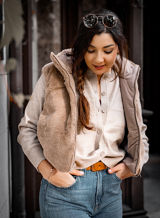 Weste aus Fake Fur Eine junge Frau steht draußen und blickt lächelnd nach unten. Sie trägt eine beige Strickjacke mit Knöpfen, eine hellbraune Weste mit flauschiger Vorderseite und Jeans mit braunem Gürtel. Ihre langen Haare sind seitlich zu einem Zopf geflochten, und auf ihrem Kopf trägt sie eine Sonnenbrille. Der Hintergrund ist leicht unscharf und zeigt eine Hausfassade.