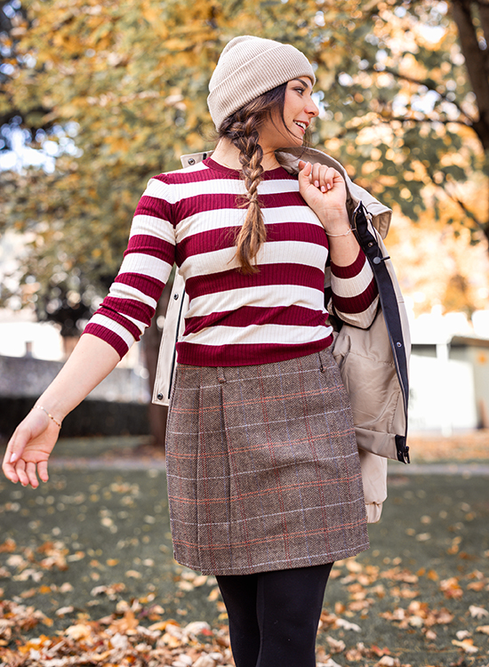 Gekonnter Mustermix Eine junge Frau steht im herbstlichen Park und lächelt, während sie sich zur Seite dreht. Sie trägt ein langärmliges Oberteil mit breiten weinroten und weißen Streifen, einen kurzen karrierten Rock in Brauntönen, schwarze Strumpfhosen und eine beige Strickhaube. Über der Schulter hat sie eine beigefarbene Jacke gelegt. Im Hintergrund sind herbstlich gefärbte Bäume und herabgefallene Blätter auf dem Boden zu sehen.