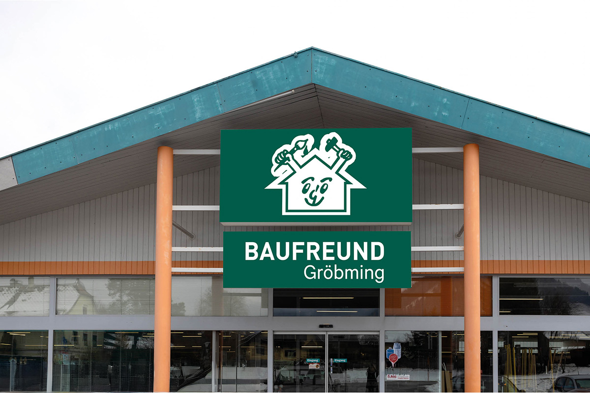 Eingang Außenansicht des neu eröffneten Landmarkt Lagerhaus Gröbming mit Glasfassade und bunten Blumenbeeten im Vordergrund. Auf der Fassade stehen die Schriftzüge „Haus & Garten“ und „Lagerhaus“. Im Hintergrund sind die beeindruckenden Berge des Ennstals zu sehen. An der Wand ist ein Plakat mit dem Slogan „Deine Lehre bei Landmarkt – Fix die beste Wahl!“ angebracht.