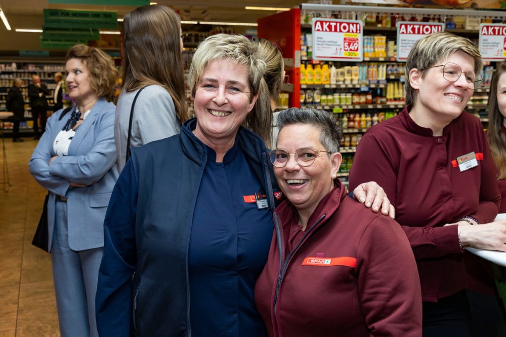 Die Marktleiterin des SPAR-Marktes Rottenmann steht gemeinsam mit einer Mitarbeiterin im Verkaufsbereich des Supermarktes und lächelt in die Kamera. Im Hintergrund sind weitere Gäste der Eröffnungsfeier sowie Regale mit Lebensmitteln zu sehen.