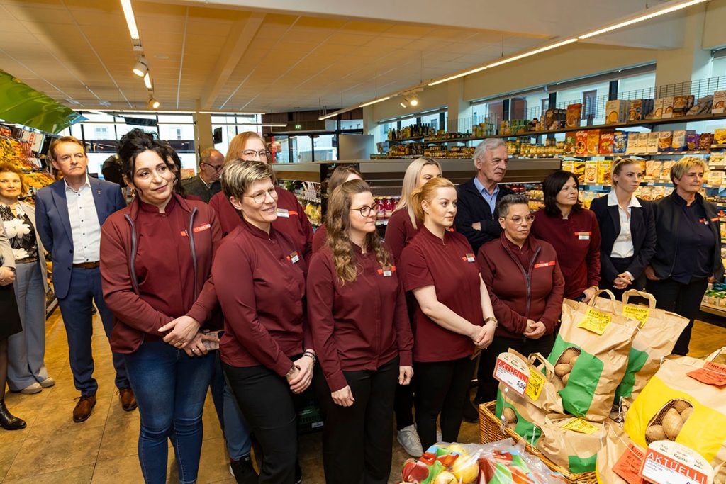 Mitarbeiterinnen des SPAR-Marktes Rottenmann sowie Gäste der Eröffnungsfeier stehen gemeinsam im Verkaufsbereich des Supermarktes und hören aufmerksam zu. Im Vordergrund sind Verkaufsstände mit Obst und Gemüse zu sehen, im Hintergrund Regale mit Lebensmitteln.